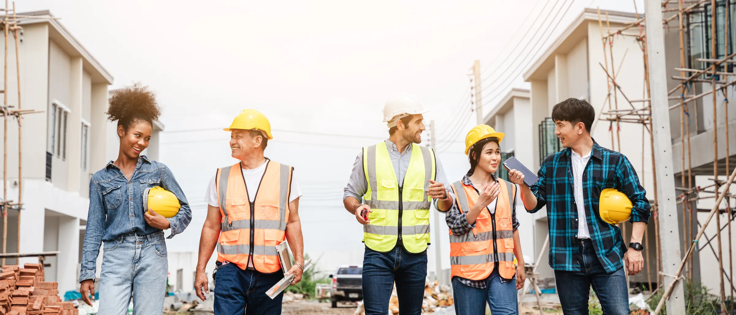 A crew of five construction workers in hard hats and hi-vis vests walking a job site together, talking and smiling — back at work